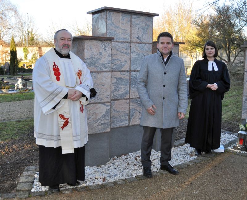 Foto ziegt Bürgermeister Holger Jung mit den beiden Geistlichen vor der Urnenwand.