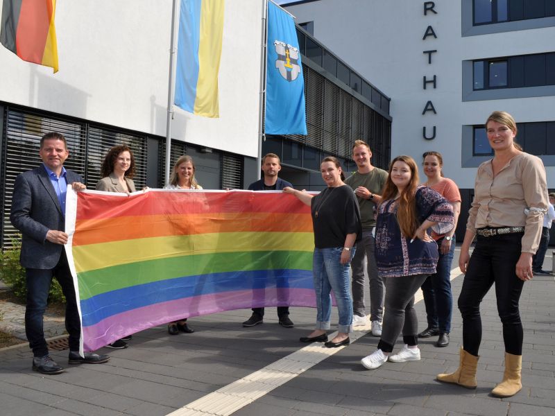 Foto zeigt Bürgermeister Holger Jung mit seinen Mitarbeitenden und der Regenbogenflagge vor dem Rathaus. 